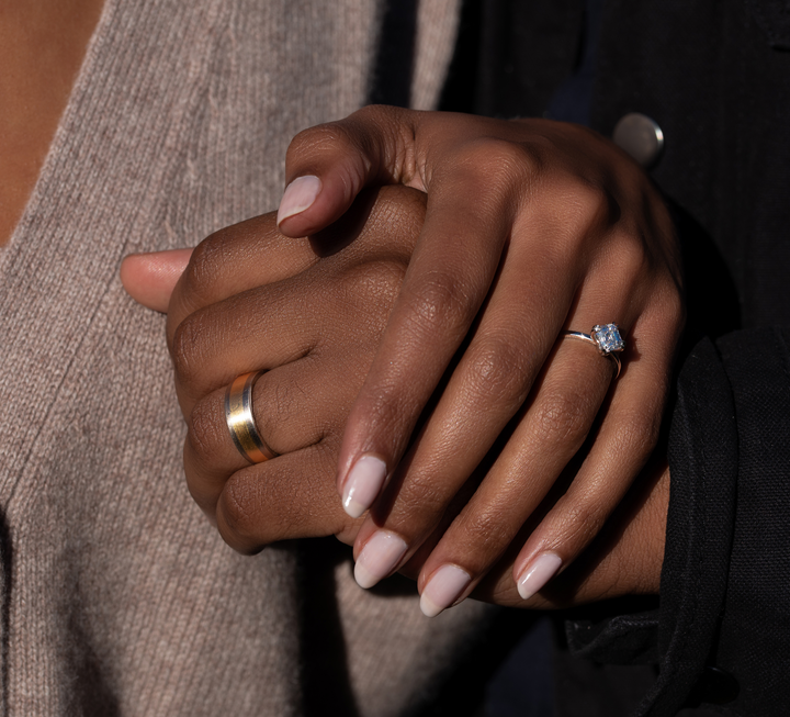 Couple embracing hands with one wearing a two toned wedding band while other wearing our Lara / White Diamond engagement ring.