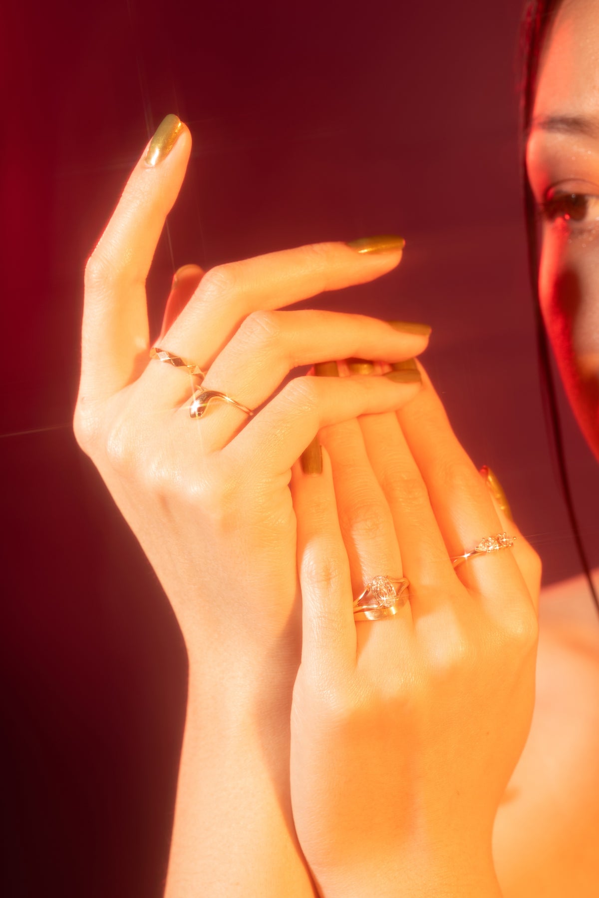 A close-up lifestyle shot of a person's hands wearing multiple delicate gold rings, with warm orange and red lighting against a dark background.